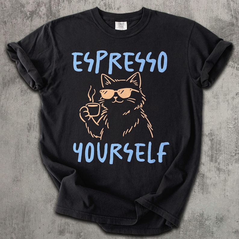 May include: Black t-shirt with the phrase "ESPRESSO YOURSELF" in light blue. A cartoon cat wearing sunglasses and holding a coffee cup is in the center. The cat is drawn in a light orange color.