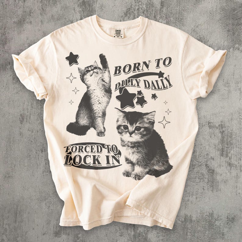 May include: Cream-coloured t-shirt with two grayscale kitten illustrations and the text "BORN TO DILLY DALLY FORCED TO LOCK IN." The design includes star graphics. The shirt is made of a soft, comfortable material.