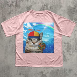 May include: A light pink t-shirt featuring a graphic of a cat wearing a colorful hat and holding a lollipop. The graphic includes a rainbow and a blue sky background. The cat has a striped pattern.