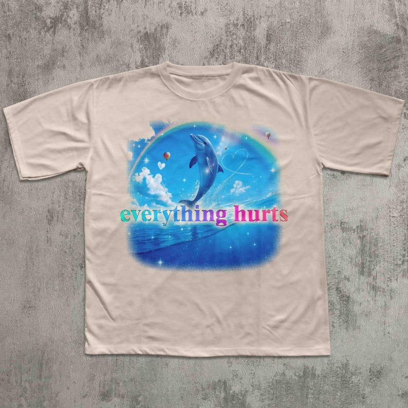 May include: Beige t-shirt featuring a vibrant graphic of a dolphin leaping from blue ocean waves under a rainbow. The text "everything hurts" is displayed in colorful letters across the bottom of the design. The shirt has short sleeves and a crew neck.