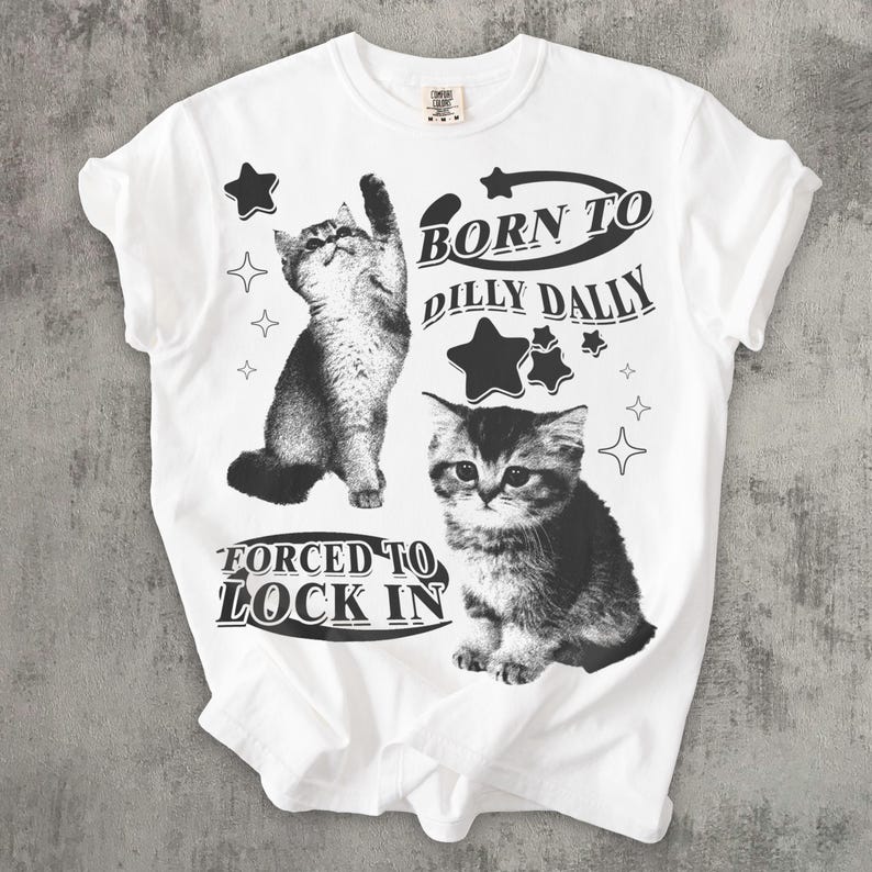 May include: White t-shirt with two grayscale cat illustrations and star graphics. The text reads "BORN TO DILLY DALLY" and "FORCED TO LOCK IN". The shirt has a crew neck and short sleeves.