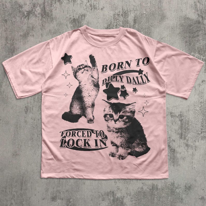 May include: A pale pink t-shirt with two black and white cat illustrations and the text "BORN TO DILLY DALLY" and "FORCED TO LOCK IN". The top has a crew neck and short sleeves.