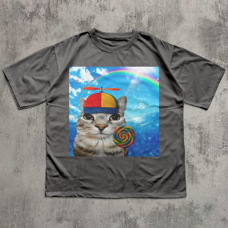 May include: Gray t-shirt featuring a whimsical graphic of a cat wearing a propeller hat and holding a colorful lollipop. The background includes a rainbow and a blue sky with clouds. The cat's expression is curious.