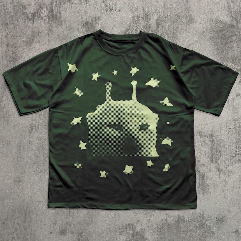 Puede incluir: Camiseta verde oscuro con una criatura extraterrestre verde claro y estrellas. El gr&aacute;fico es un dise&ntilde;o de semitonos. La camiseta tiene mangas cortas y cuello redondo.