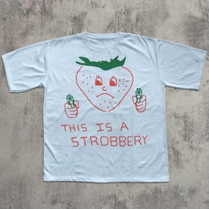 May include: Light blue t-shirt with a red strawberry graphic holding two green guns. The strawberry has a sad face and the text "THIS IS A STROBBERY" is printed below the graphic.