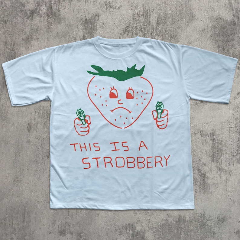 May include: Light blue t-shirt with a red strawberry graphic holding two green guns. The strawberry has a sad face and the text "THIS IS A STROBBERY" is printed below the graphic.