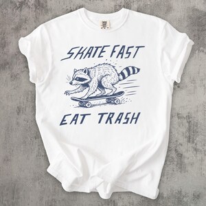 May include: White t-shirt with a blue graphic of a raccoon skateboarding. The text "SKATE FAST EAT TRASH" is above and below the raccoon illustration. The shirt has a crew neck and short sleeves.