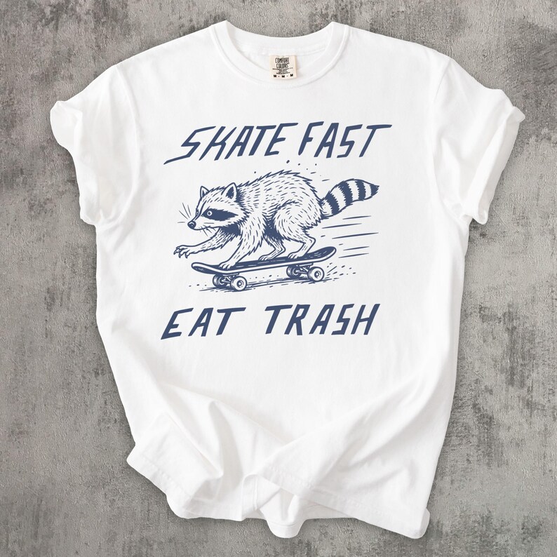 May include: White t-shirt with a blue graphic of a raccoon skateboarding. The text "SKATE FAST EAT TRASH" is above and below the raccoon illustration. The shirt has a crew neck and short sleeves.