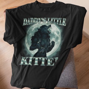 Pode incluir: Camiseta preta com uma ilustra&ccedil;&atilde;o de lobisomem em frente a uma lua cheia. O texto "DADDY'S LITTLE KITTEN" &eacute; impresso acima e abaixo da imagem em uma fonte prateada estilizada. O design &eacute; centralizado na camiseta.