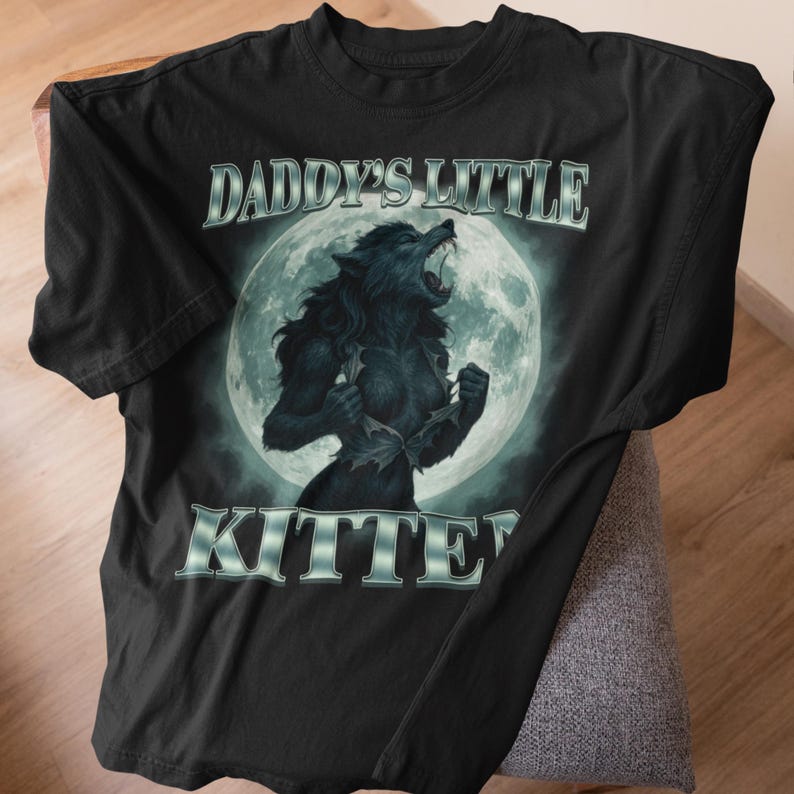 Pode incluir: Camiseta preta com uma ilustra&ccedil;&atilde;o de lobisomem em frente a uma lua cheia. O texto "DADDY'S LITTLE KITTEN" &eacute; impresso acima e abaixo da imagem em uma fonte prateada estilizada. O design &eacute; centralizado na camiseta.