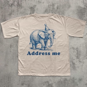 May include: Cream-colored t-shirt featuring a blue illustration of an elephant wearing a party hat. The text "Address me" is printed in blue below the elephant. The shirt has short sleeves and a crew neck.