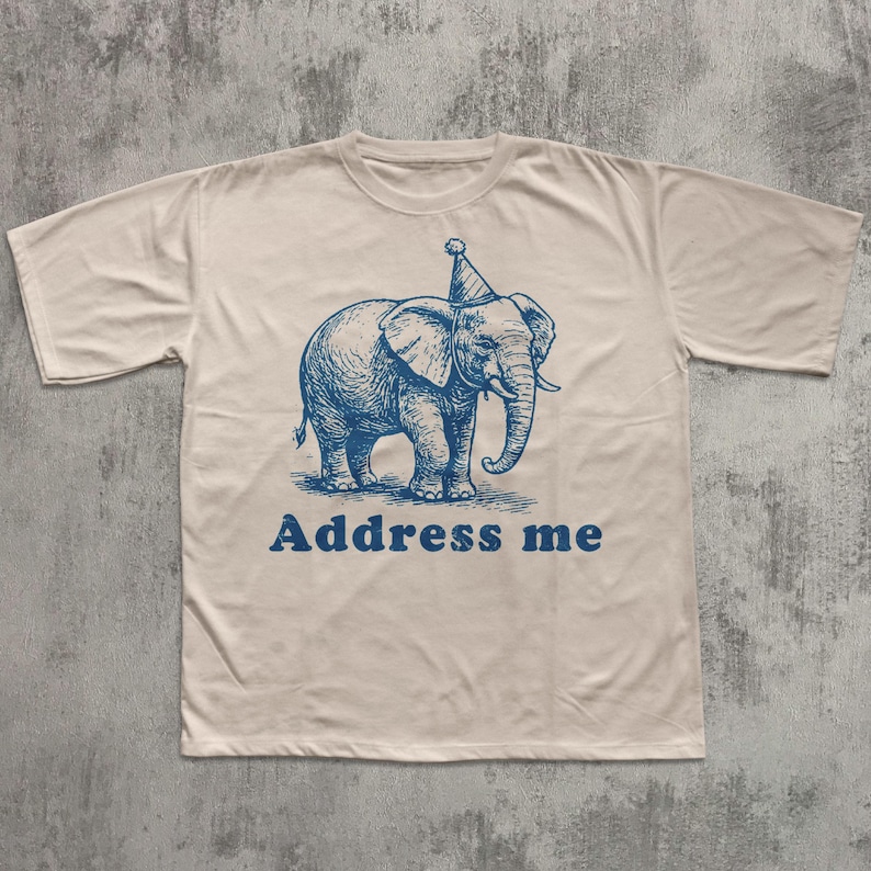 May include: Cream-colored t-shirt featuring a blue illustration of an elephant wearing a party hat. The text "Address me" is printed in blue below the elephant. The shirt has short sleeves and a crew neck.