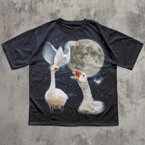 Op de afbeelding: Een zwart t-shirt met korte mouwen en een grafische print van drie witte ganzen tegen een donkere nachtelijke hemel. E&eacute;n gans vliegt, &eacute;&eacute;n staat en &eacute;&eacute;n gans snatert met de snavel open, allemaal onder een grote volle maan.