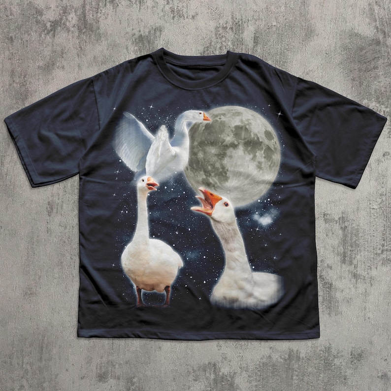 Op de afbeelding: Een zwart t-shirt met korte mouwen en een grafische print van drie witte ganzen tegen een donkere nachtelijke hemel. E&eacute;n gans vliegt, &eacute;&eacute;n staat en &eacute;&eacute;n gans snatert met de snavel open, allemaal onder een grote volle maan.
