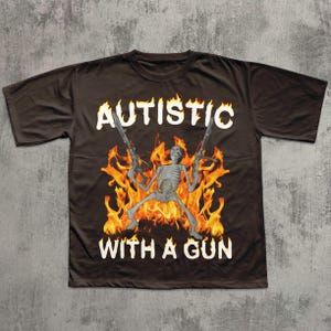 May include: Dark brown t-shirt with the words "AUTISTIC" and "WITH A GUN" in white. The graphic features a skeleton figure holding two guns, set against a backdrop of flames.