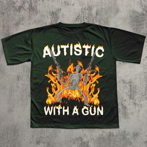 May include: Dark green t-shirt with the text "AUTISTIC WITH A GUN" in white, featuring a skeleton holding two guns in front of a fiery background. The shirt is a crew neck style.