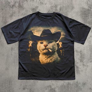 May include: Navy blue t-shirt featuring a cat wearing a cowboy hat and smoking a cigarette. The graphic includes cows and a mountain range in the background. The cat has a squinting expression. The shirt is a casual wear item.