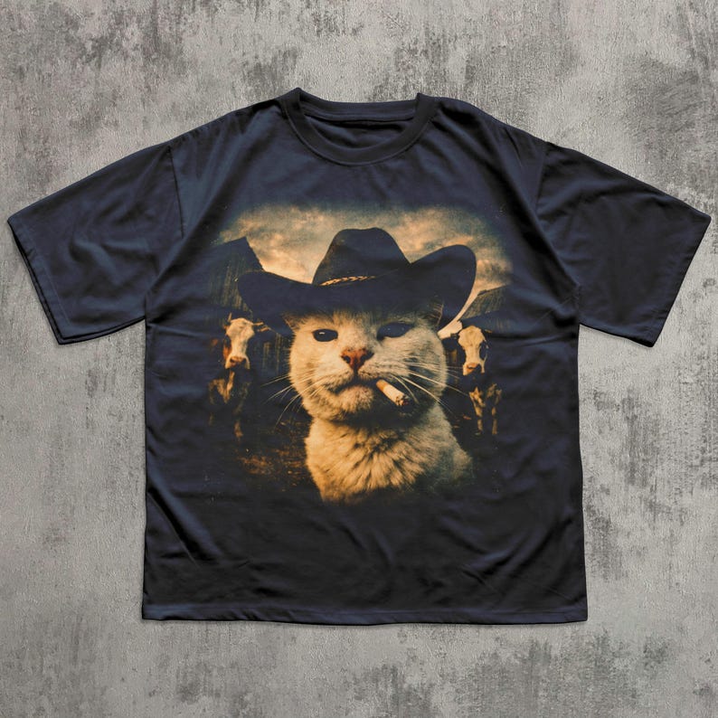 May include: Navy blue t-shirt featuring a cat wearing a cowboy hat and smoking a cigarette. The graphic includes cows and a mountain range in the background. The cat has a squinting expression. The shirt is a casual wear item.