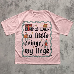 Puede incluir: Una camiseta de manga corta de color rosa claro con un gr&aacute;fico central rodeado por un borde de enredadera verde. El dise&ntilde;o incluye dos caballeros, un cofre del tesoro, estrellas y el texto negro "That was a little cringe, my liege" con una T roja.