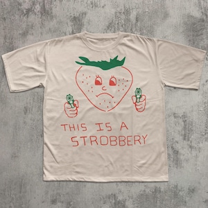 May include: Off-white t-shirt with a red and green graphic of a strawberry holding two guns. The strawberry has a sad face and the text "THIS IS A STROBBERY" is printed below the graphic.