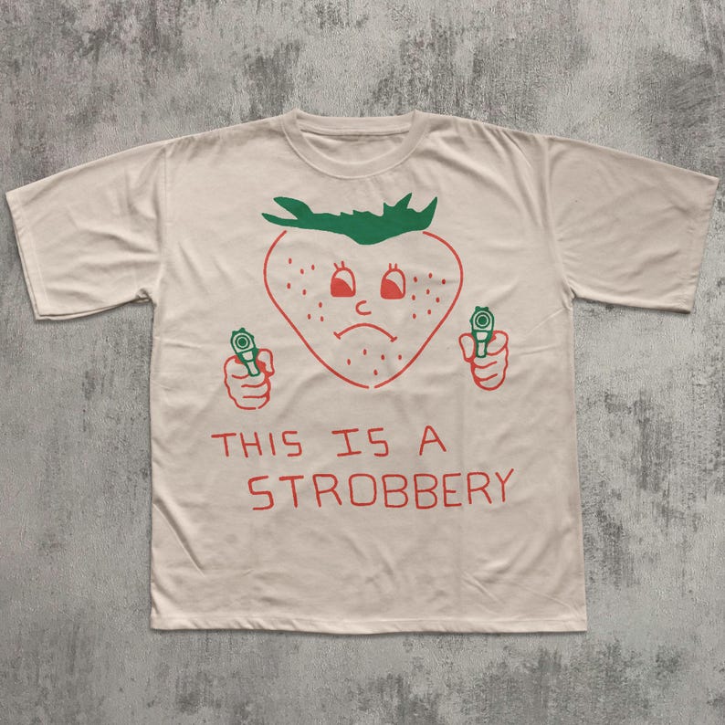 May include: Off-white t-shirt with a red and green graphic of a strawberry holding two guns. The strawberry has a sad face and the text "THIS IS A STROBBERY" is printed below the graphic.