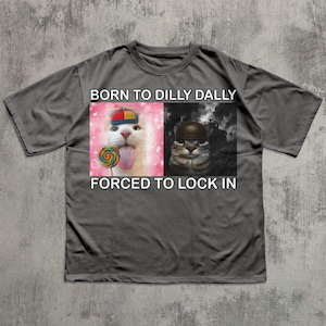 May include: Gray t-shirt with the text "BORN TO DILLY DALLY FORCED TO LOCK IN". The shirt features a split image of two cats. One cat is wearing a hat and holding a lollipop, and the other is wearing a helmet.