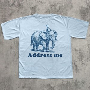 May include: Light blue t-shirt featuring a blue illustration of an elephant wearing a party hat. The text "Address me" is printed below the elephant in a matching blue color. The shirt has short sleeves and a crew neck.