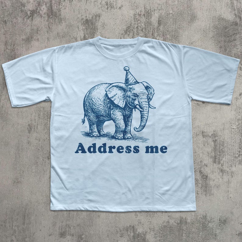 May include: Light blue t-shirt featuring a blue illustration of an elephant wearing a party hat. The text "Address me" is printed below the elephant in a matching blue color. The shirt has short sleeves and a crew neck.