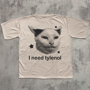 May include: A cream-colored t-shirt featuring a black and white image of a cat's face with three black stars. Below the cat image, the text reads "I need tylenol".