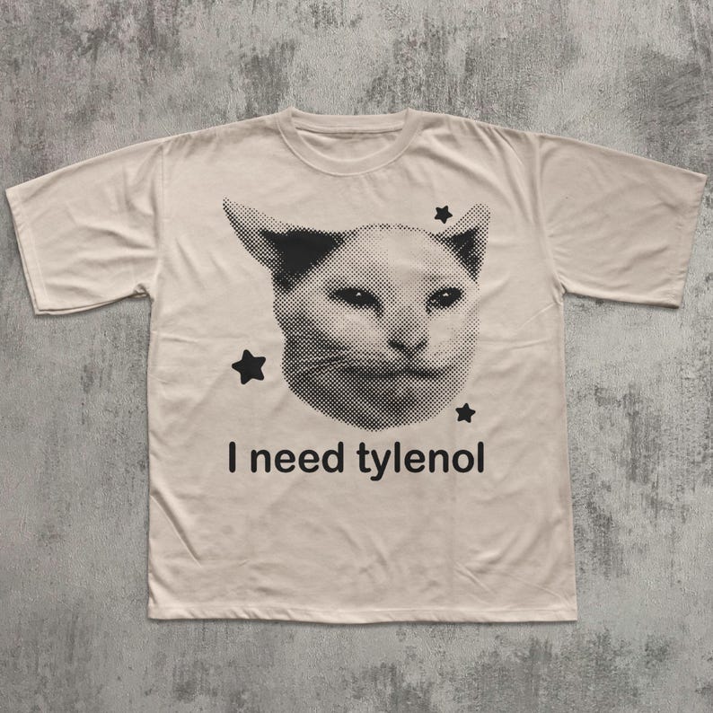 May include: A cream-colored t-shirt featuring a black and white image of a cat's face with three black stars. Below the cat image, the text reads "I need tylenol".