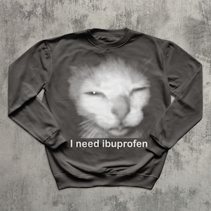 May include: Dark gray sweatshirt featuring a white halftone image of a cat's face. The text "I need ibuprofen" is printed in white below the image. The sweatshirt has long sleeves and a crew neck.