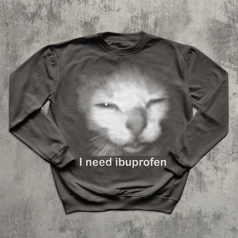 May include: Dark gray sweatshirt featuring a white halftone image of a cat's face. The text "I need ibuprofen" is printed in white below the image. The sweatshirt has long sleeves and a crew neck.