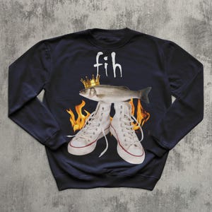 May include: Navy blue sweatshirt with a graphic design. It features a fish wearing a gold crown above white high-top trainers. The word "fih" is written above the fish in white. Flames are visible behind the trainers.