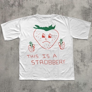 May include: White t-shirt with a red and green graphic of a strawberry holding two guns. The strawberry has a sad face and the text "THIS IS A STROBBERY" is printed below the graphic.