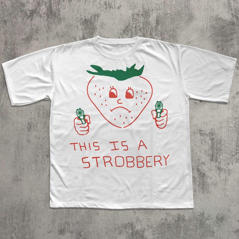 May include: White t-shirt with a red and green graphic of a strawberry holding two guns. The strawberry has a sad face and the text "THIS IS A STROBBERY" is printed below the graphic.