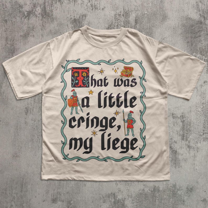 Puede incluir: Una camiseta beige de manga corta con un gr&aacute;fico rodeado por un borde de enredadera color verde azulado. El texto "That was a little cringe, my liege" aparece en fuente g&oacute;tica negra, junto a una letra T roja, dos caballeros, un cofre y estrellas.
