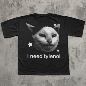 May include: Black t-shirt featuring a white, pixelated cat face with three white stars. The text "I need tylenol" is printed in white below the cat image. The shirt is made of a soft material.