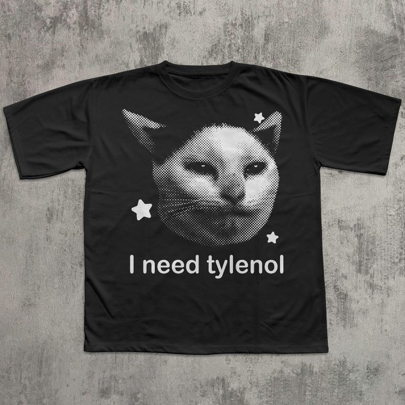 May include: Black t-shirt featuring a white, pixelated cat face with three white stars. The text "I need tylenol" is printed in white below the cat image. The shirt is made of a soft material.