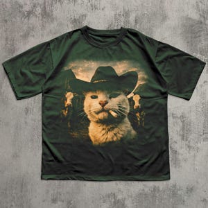 May include: Dark green t-shirt featuring a graphic of a cat wearing a cowboy hat and smoking a cigarette. The cat is set against a backdrop of cows and mountains. The shirt is a relaxed fit.