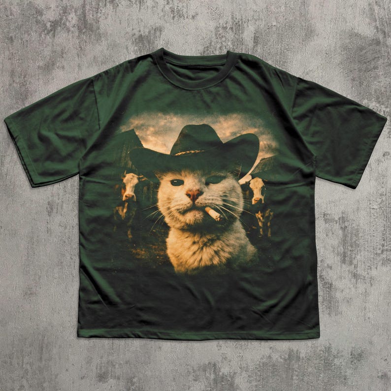May include: Dark green t-shirt featuring a graphic of a cat wearing a cowboy hat and smoking a cigarette. The cat is set against a backdrop of cows and mountains. The shirt is a relaxed fit.