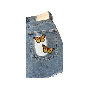 May include: A pair of denim shorts with a back pocket featuring two embroidered butterflies. The butterflies are orange and black with white accents.