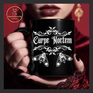 Op de afbeelding: Zwarte keramische mok met de witte tekst "Carpe Noctem" en motillustraties. De mok wordt vastgehouden door een persoon met zwarte nagellak. De mok heeft een zwart handvat.