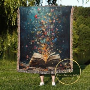 May include: A woven tapestry throw blanket with a dark blue background featuring a book with colorful butterflies flying out of it. The book is open and the pages are textured. The butterflies are in various shades of blue, purple, pink, yellow, and orange.