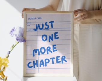 May include: A framed print with the text "JUST ONE MORE CHAPTER" in blue on a pink library card design. The frame is light wood. A sprig of purple and yellow flowers is in the corner.