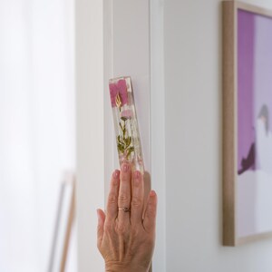 K&ouml;nnte beinhalten: Ein rechteckiger, klarer Mezuzah-Kasten aus Harz mit getrockneten rosa Blumen und goldenen Akzenten. Der Kasten ist an einem wei&szlig;en T&uuml;rrahmen befestigt. Eine Hand mit einem Ring ber&uuml;hrt die Mezuzah.