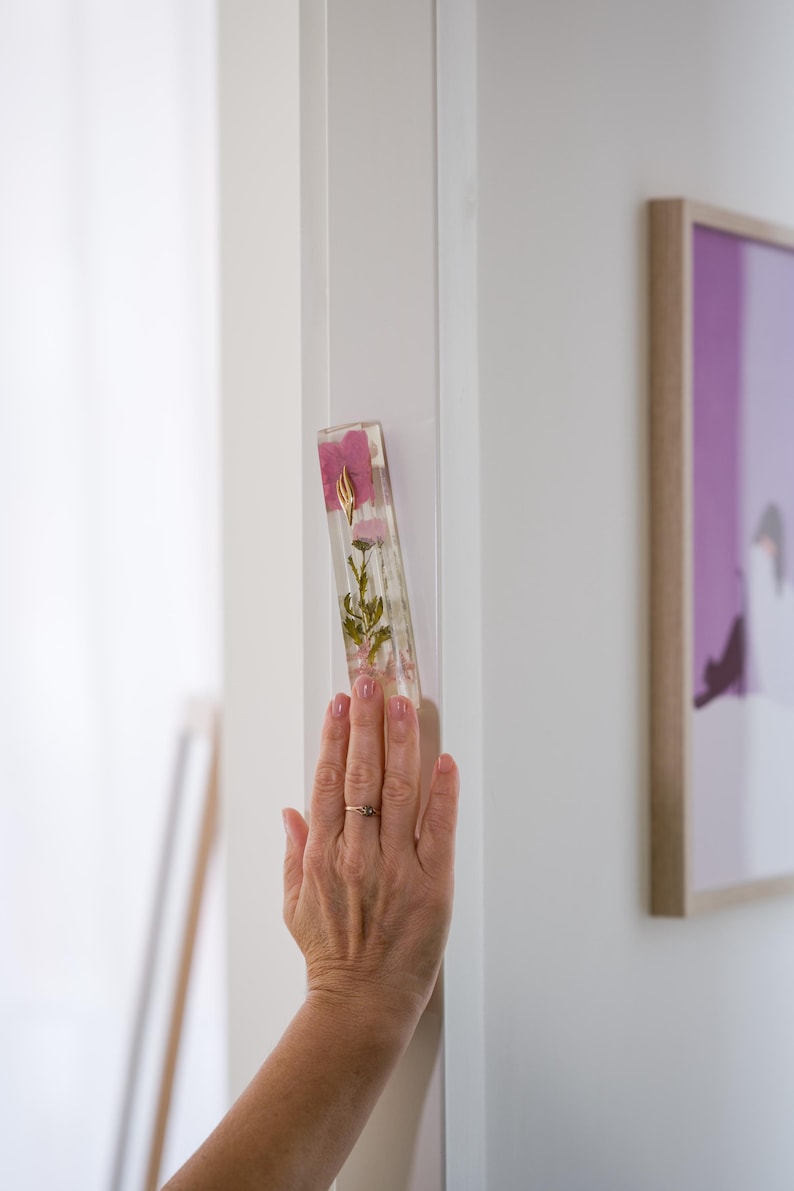 K&ouml;nnte beinhalten: Ein rechteckiger, klarer Mezuzah-Kasten aus Harz mit getrockneten rosa Blumen und goldenen Akzenten. Der Kasten ist an einem wei&szlig;en T&uuml;rrahmen befestigt. Eine Hand mit einem Ring ber&uuml;hrt die Mezuzah.