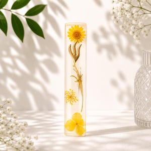 K&ouml;nnte beinhalten: Ein klarer, rechteckiger Harzblock mit einer gelben Blume und anderen getrockneten floralen Elementen im Inneren. Ein goldfarbenes Dekorationselement ist ebenfalls eingebettet. Der Hintergrund ist wei&szlig; mit weichen Schatten.