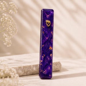 May include: A rectangular, purple mezuzah case with gold leaf accents and a gold-coloured Hebrew letter. The case is displayed on a light-coloured stone surface, with white flowers in the background.