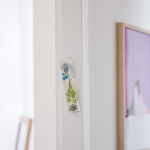 K&ouml;nnte beinhalten: Ein rechteckiger, klarer Harzmagnet mit eingebetteten getrockneten Blumen und Bl&auml;ttern. Der Magnet ist an einem wei&szlig;en T&uuml;rrahmen befestigt. Die botanischen Elemente umfassen blaue Blumen, gr&uuml;ne Farne und kleine braune Bl&auml;tter.
