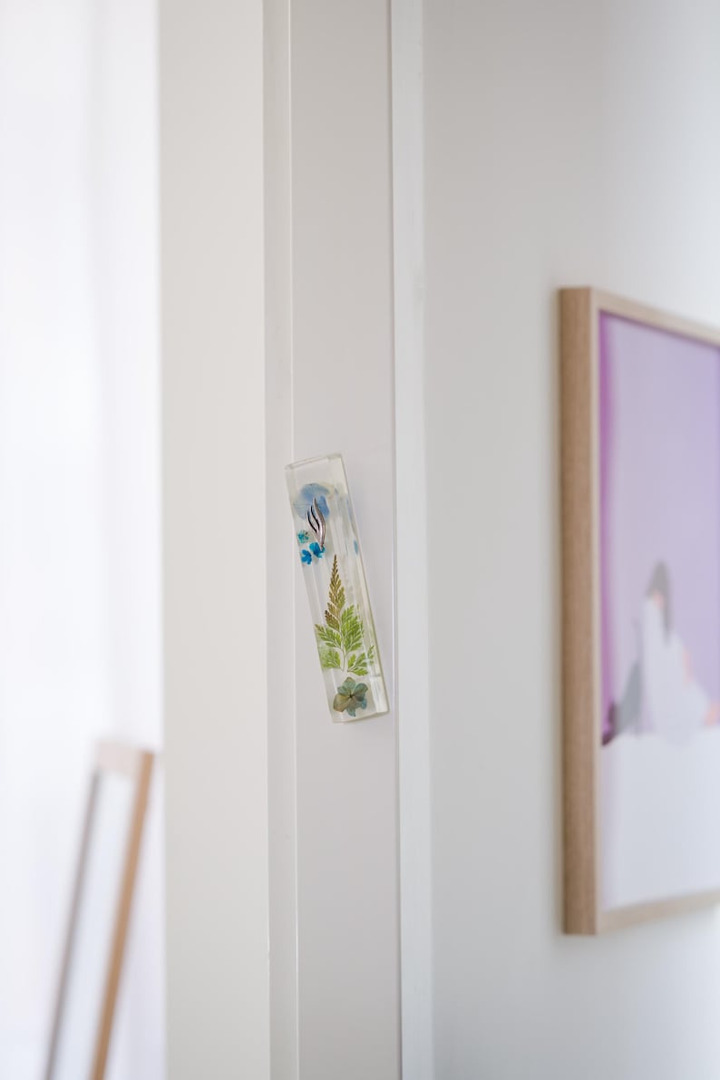 K&ouml;nnte beinhalten: Ein rechteckiger, klarer Harzmagnet mit eingebetteten getrockneten Blumen und Bl&auml;ttern. Der Magnet ist an einem wei&szlig;en T&uuml;rrahmen befestigt. Die botanischen Elemente umfassen blaue Blumen, gr&uuml;ne Farne und kleine braune Bl&auml;tter.